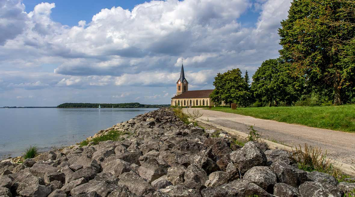 Grey rocks stretching along the side of a blue lake on the left and a grey path with a spire building in the background and green grass and trees